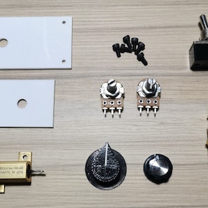 May include: An assortment of electronic components, including white plastic panels, a toggle switch, potentiometers, a brass fitting, and a resistor. The components are arranged on a light wood-grain surface, suitable for DIY electronics projects.