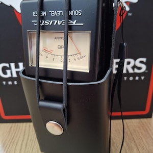 May include: A black Realistic sound level meter in a black leather case. The meter has a clear display with a needle and markings for dB levels. The case has a strap and a snap closure. The text "REALISTIC" and "SOUND LEVEL METER" are visible.