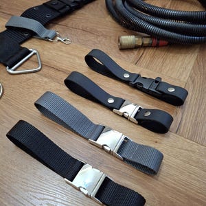 Puede incluir: Un conjunto de correas negras y grises con hebillas plateadas. Las correas están hechas de cinta y tienen diferentes longitudes. También hay una manguera negra con una franja roja y un conector de latón.