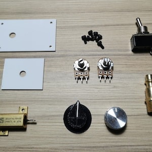 May include: An assortment of electronic components, including two white metal plates, a black toggle switch, two potentiometers, a brass fitting, a resistor, a black knob, a silver knob, and black screws. The components are arranged on a light brown wooden surface.