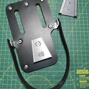 May include: A black leather holster with a silver metal plate and a black strap. The holster is designed to hold a small object, such as a knife or a tool.