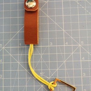May include: A brown leather key fob with a silver press stud. It features a yellow cord connected to a gold-coloured rectangular metal tag. The background is a blue grid.