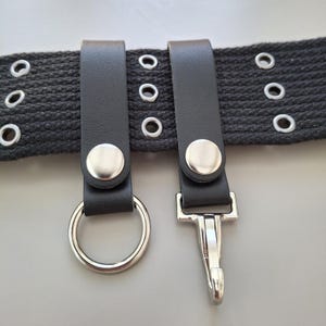 Può includere: Una cintura in tessuto nero intrecciato con tre file di occhielli in metallo argentato. Due passanti in similpelle nera sono fissati alla cintura con bottoni a pressione argentati. Un passante regge un anello metallico, l'altro un moschettone.