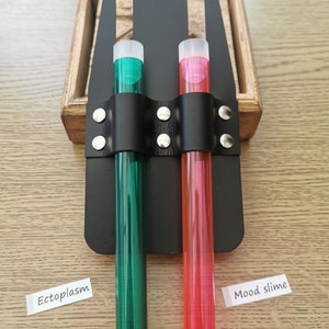 Könnte beinhalten: Zwei durchsichtige Plastikröhrchen mit Deckeln, eines grün und eines rosa, werden in einem schwarzen Lederhalter mit silbernen Nieten gehalten. Die Röhrchen sind mit "Ectoplasm" und "Mood slime" beschriftet.