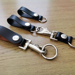 Peut inclure: Trois porte-clés en cuir noir avec des fermoirs et des anneaux en métal argenté. Les porte-clés ont une fermeture à bouton.