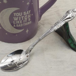 K&ouml;nnte beinhalten: Ein silberner L&ouml;ffel mit aufwendigen Details und einem Halbmondsymbol auf der Sch&uuml;ssel. Der L&ouml;ffel liegt auf einem gr&uuml;nen St&auml;nder neben einer lila Tasse mit dem Text "YOU SAY WITCH LIKE IT'S A BAD THING."