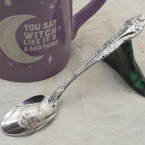 May include: A silver spoon with an engraved pentagram design on the bowl, resting on a green stand. A purple mug with the text "YOU SAY WITCH LIKE IT'S A BAD THING" is in the background. The spoon has ornate detailing.