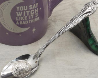 May include: A silver spoon with an engraved pentagram design on the bowl, resting on a green stand. A purple mug with the text "YOU SAY WITCH LIKE IT'S A BAD THING" is in the background. The spoon has ornate detailing.
