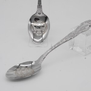 May include: Two silver spoons with ornate floral and scrollwork designs. One spoon is in the foreground, and another is in the background. The spoons have a reflective surface and are likely made of sterling silver or a similar metal.