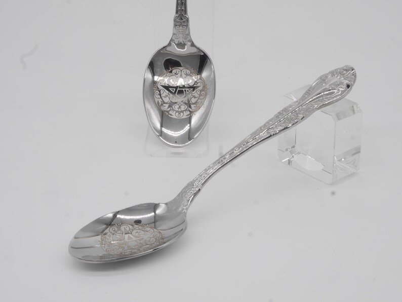 May include: Two silver spoons with ornate floral and scrollwork designs. One spoon is in the foreground, and another is in the background. The spoons have a reflective surface and are likely made of sterling silver or a similar metal.