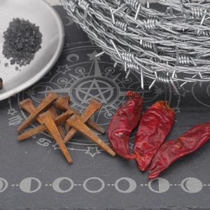 May include: A still life arrangement featuring a silver plate with black peppercorns and dark salt, rusty nails, and three dried red chili peppers. A coil of barbed wire sits in the background on a dark surface with a celestial design.
