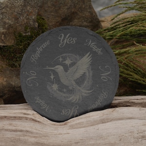 May include: A black slate coaster with a hummingbird design and the words "Yes", "Maybe", "No", and "Rephrase" written around the edge.