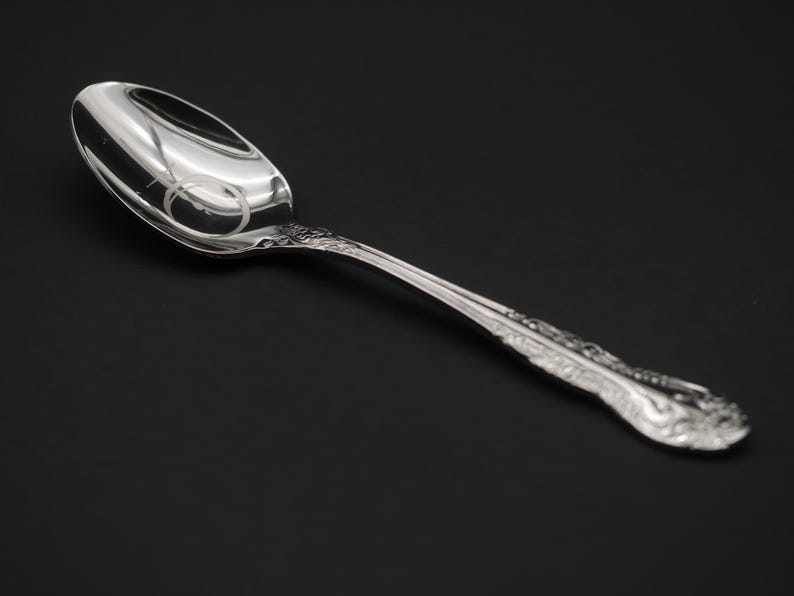 May include: A silver spoon with an oval bowl and a decorative handle. The spoon is highly polished, reflecting the surrounding environment. The handle features an ornate floral design. The spoon is set against a dark background.