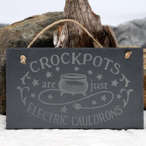 May include: A black slate sign with white text that reads "Crockpots are just electric cauldrons". The sign has a rope hanger and a stylized image of a cauldron with stars around it.