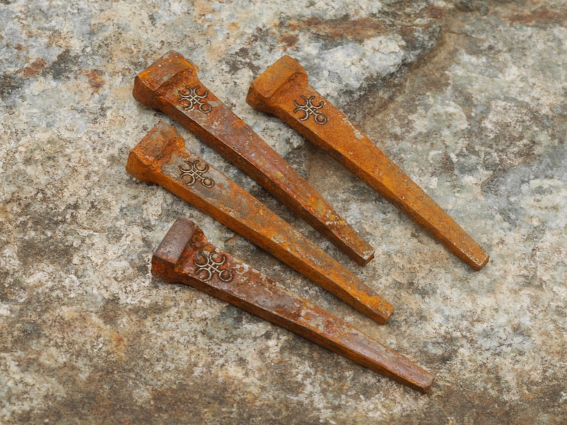 May include: Four rusty, antique-style horseshoe nails with decorative floral designs on the heads. The nails are a warm, earthy brown color and are arranged on a textured, stone-like surface.