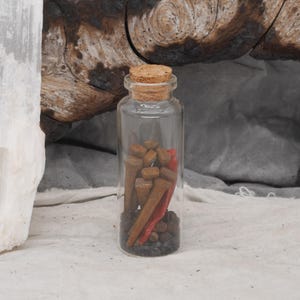 May include: A clear glass bottle with a cork stopper, filled with small brown objects, a red item, and dark gray pebbles. The bottle is set against a neutral background with a piece of wood and a white stone.