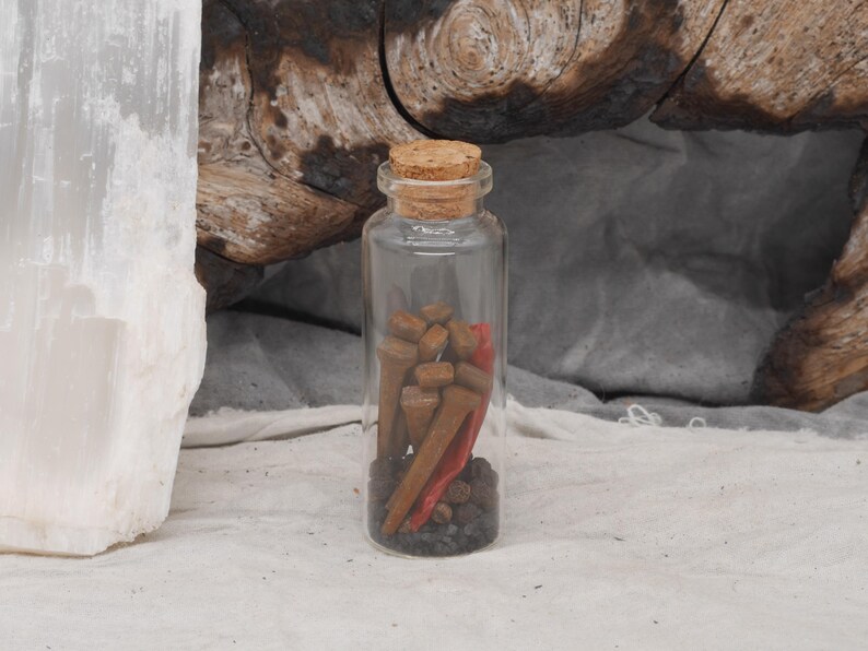 May include: A clear glass bottle with a cork stopper, filled with small brown objects, a red item, and dark gray pebbles. The bottle is set against a neutral background with a piece of wood and a white stone.