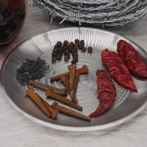 May include: A silver plate holds various items: black peppercorns, dark gray granules, rusty nails, and three dried red chili peppers. A glass bottle and barbed wire are in the background.