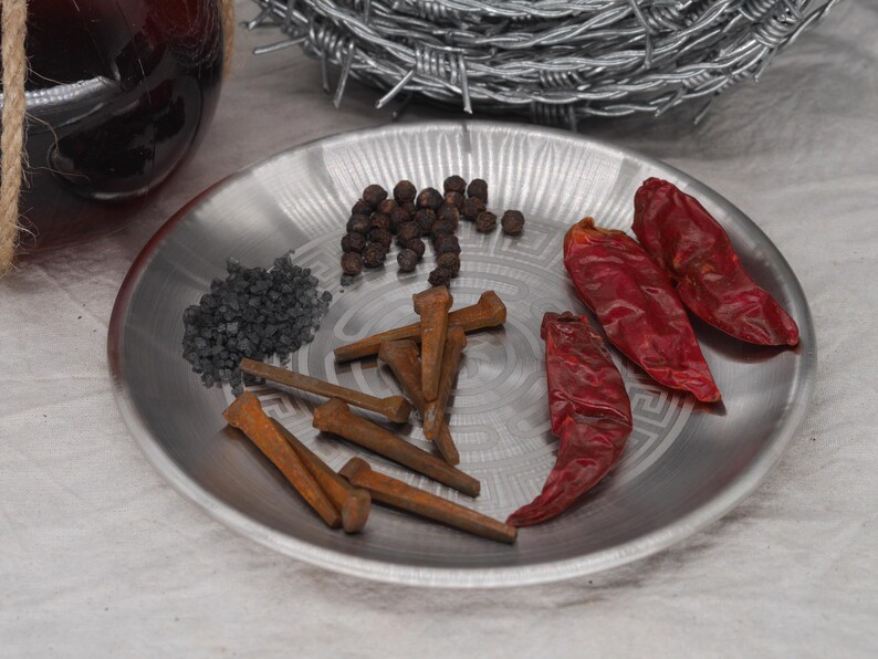 May include: A silver plate holds various items: black peppercorns, dark gray granules, rusty nails, and three dried red chili peppers. A glass bottle and barbed wire are in the background.