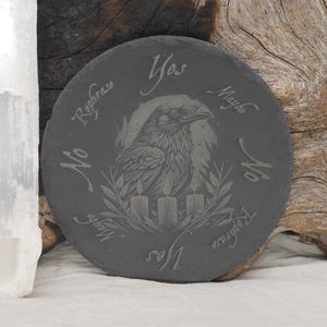 May include: Round, dark gray slate coaster with an engraved raven design. The raven is surrounded by the words "Yes", "Maybe", "No", and "Rephrase". Three candles and a laurel wreath are also engraved on the coaster.