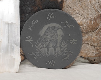 May include: Circular, dark grey slate coaster featuring an engraved raven design. The coaster has the words "Yes", "Maybe", "No", and "Rephrase" around the raven. Three candles and a laurel wreath are also engraved.