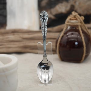 May include: A decorative silver spoon with an intricate handle design, presented on a clear stand. The spoon's bowl is curved and reflective. A dark glass bottle, wrapped with rope, is visible in the background.