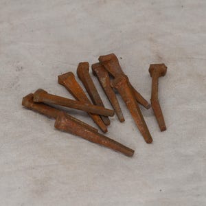 May include: A collection of rusty, antique nails with rectangular heads and tapered bodies. The nails are various sizes and are scattered on a light-colored, textured surface. The nails have a weathered, aged appearance.