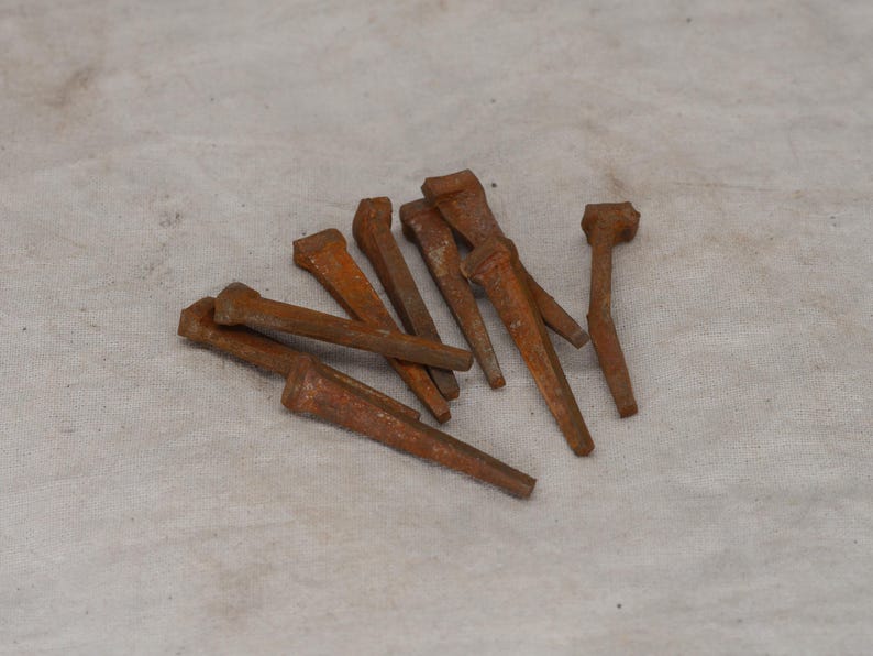 May include: A collection of rusty, antique nails with rectangular heads and tapered bodies. The nails are various sizes and are scattered on a light-colored, textured surface. The nails have a weathered, aged appearance.