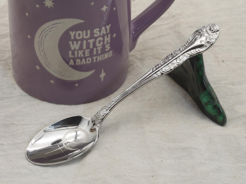 May include: A silver spoon with decorative patterns is placed on a green and black stand. A purple mug with the text "YOU SAY WITCH LIKE IT'S A BAD THING" is in the background. The spoon has a crescent moon and cross symbol.