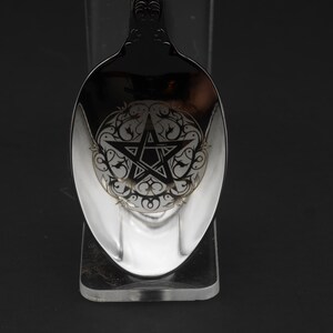 May include: A silver spoon with an ornate handle and a detailed design on the bowl. The bowl features a pentagram within a decorative circular pattern. The spoon is displayed against a dark background.