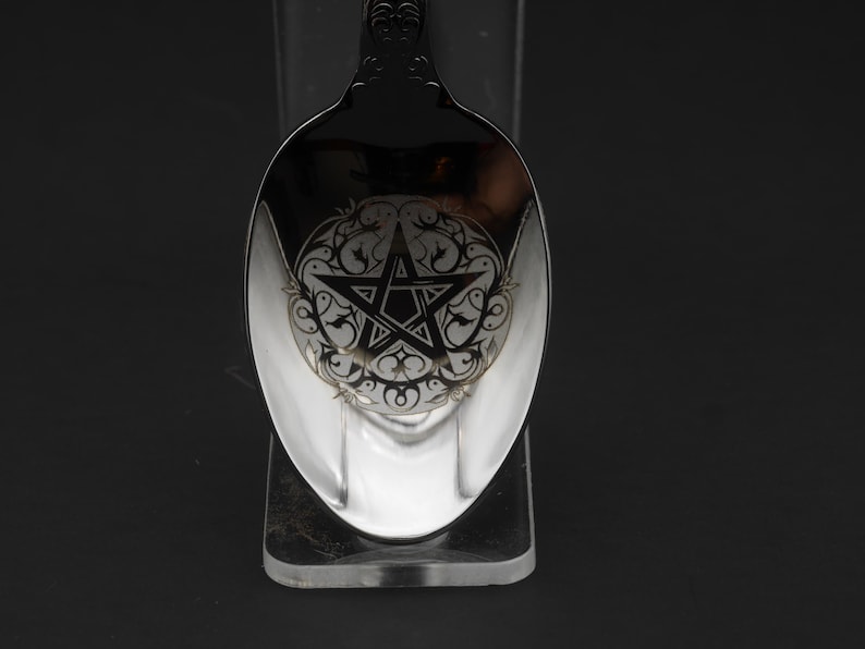 May include: A silver spoon with an ornate handle and a detailed design on the bowl. The bowl features a pentagram within a decorative circular pattern. The spoon is displayed against a dark background.
