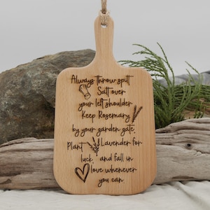 May include: A wooden cutting board with a handle, featuring engraved text and illustrations. The text includes phrases like "Always throw salt over your left shoulder" and "Plant lavender for luck." A heart and botanical illustrations are also present.