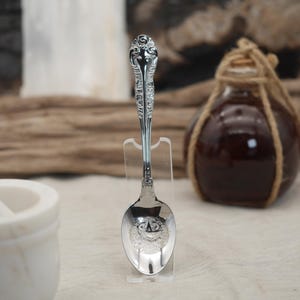 May include: A silver spoon with ornate detailing on the handle and bowl, displayed on a clear stand. The spoon's bowl features a detailed design. A dark glass bottle with a rope tied around the neck is in the background.