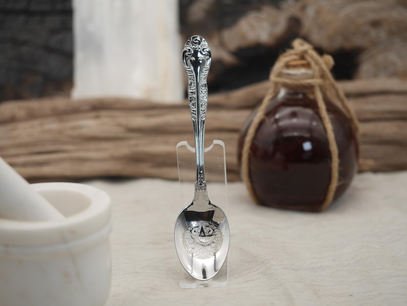 May include: A silver spoon with ornate detailing on the handle and bowl, displayed on a clear stand. The spoon's bowl features a detailed design. A dark glass bottle with a rope tied around the neck is in the background.