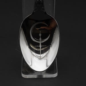 May include: A silver spoon with an engraved crescent moon and cross design. The spoon's reflective surface is set against a dark background, emphasizing the intricate details of the design. The spoon is a decorative item.
