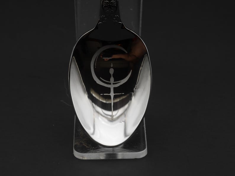 May include: A silver spoon with an engraved crescent moon and cross design. The spoon's reflective surface is set against a dark background, emphasizing the intricate details of the design. The spoon is a decorative item.