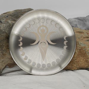 May include: A silver metal plate with a design of a stylized female figure with three crescent moons and a circle of smaller moons around the outside.