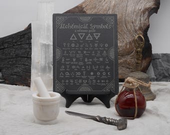 May include: A dark gray slate sign with the text "Alchemical Symbols: a reference guide" and various symbols. A white mortar and pestle, a small glass bottle with red liquid, and a twisted metal knife are in the foreground.