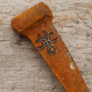 May include: Close-up of a rusty metal object with a decorative design. The object has a rectangular shape with a wider top and a detailed, dark-colored emblem in the center. The surface shows signs of age and wear.