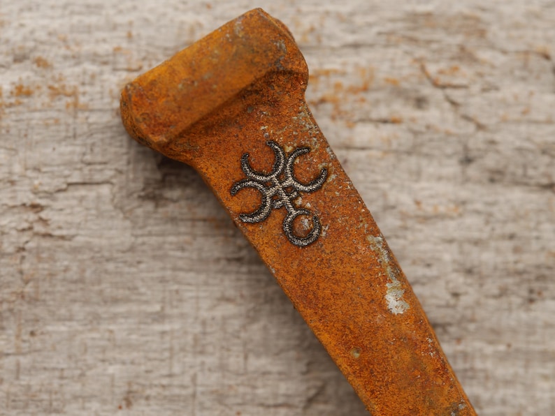May include: Close-up of a rusty metal object with a decorative design. The object has a rectangular shape with a wider top and a detailed, dark-colored emblem in the center. The surface shows signs of age and wear.