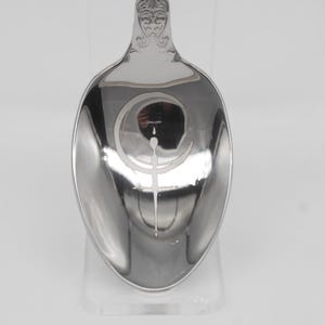 May include: A silver spoon with an engraved design inside the bowl. The design features a crescent moon and a vertical line with a small circle at the top. The handle has an ornate, decorative pattern.