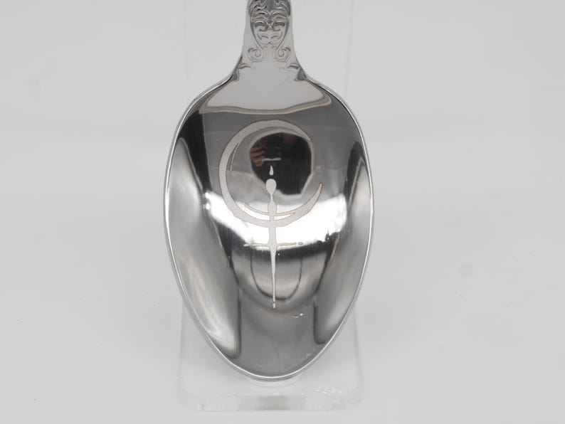 May include: A silver spoon with an engraved design inside the bowl. The design features a crescent moon and a vertical line with a small circle at the top. The handle has an ornate, decorative pattern.