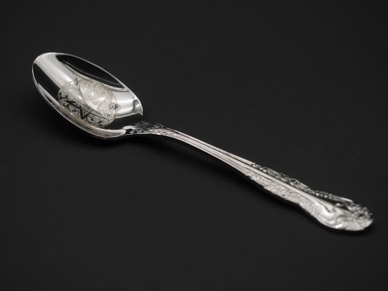 May include: A silver spoon with an ornate handle and a reflective bowl. The spoon is angled, showcasing its polished surface and intricate design. The background is a solid black.