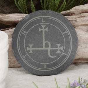 May include: A black slate coaster with a white sigil of the demon Lilith. The sigil is a cross with a serpent wrapped around it. The words "L.I.H.I.L.I." are inscribed around the sigil.