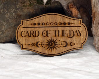 May include: A wooden sign with the text "Card of the Day" in a decorative font. The sign is decorated with a sun, moon, and stars.