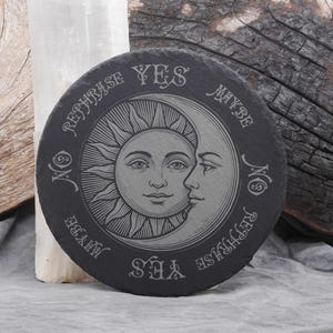 May include: A round, black slate coaster with an engraved sun and moon design. The words "YES", "NO", "MAYBE", and "REPHRASE" are etched around the edge. The coaster is a decorative item.