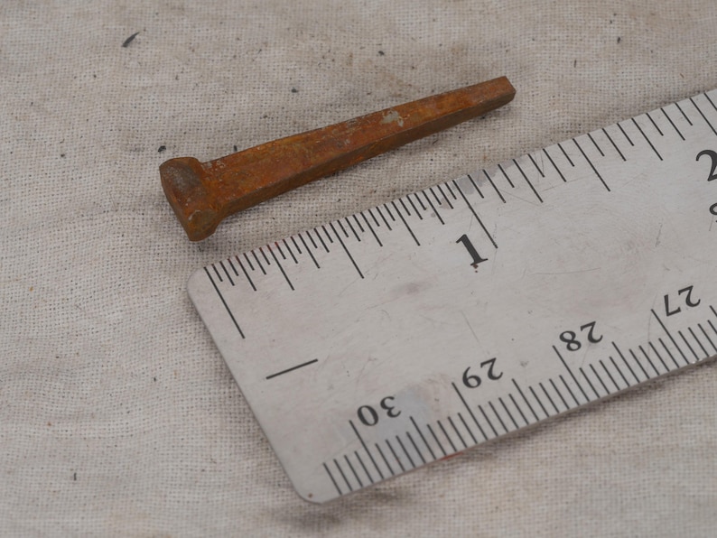 May include: A rusty, rectangular nail with a flattened head, approximately 1 inch long, lies next to a ruler. The nail's color is a reddish-brown, indicating oxidation. The ruler shows inch and fractional inch measurements.