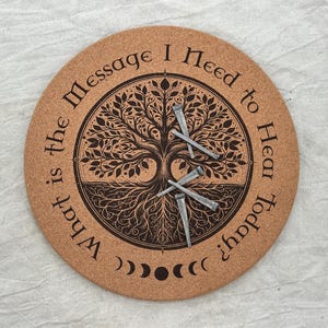 May include: Round cork coaster with a tree of life design and the text "What is the Message I Need to Hear Today?" Three silver nails are placed on the coaster. The design includes phases of the moon.