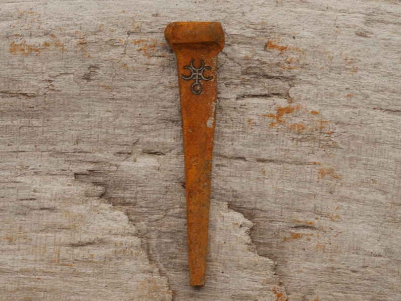 May include: A rusty, antique metal nail with a decorative emblem. The nail has a rectangular head and a long, tapered body. The emblem is a stylized design. The nail is set against a weathered wooden background.