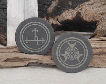 Könnte beinhalten: Zwei runde, dunkelgraue Schieferuntersetzer mit geätzten weißen Designs. Einer zeigt ein kreuzartiges Symbol in einem Kreis, der andere ein Dreifachmond- und Labyrinth-Design. Die Untersetzer werden auf einem Stück Treibholz präsentiert.